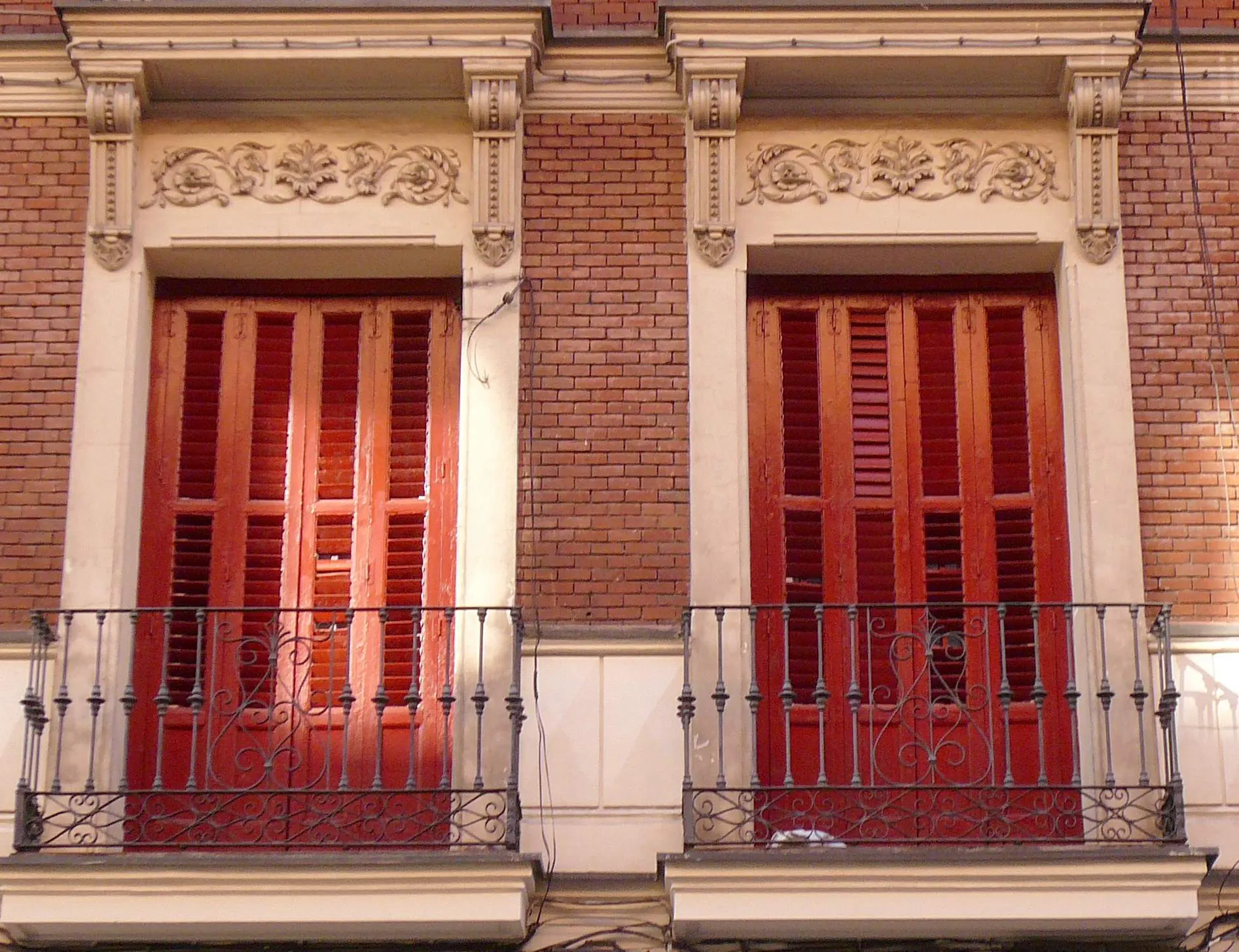 Dos contraventanas rojas tras balcones de hierro en una fachada de ladrillo de Madrid